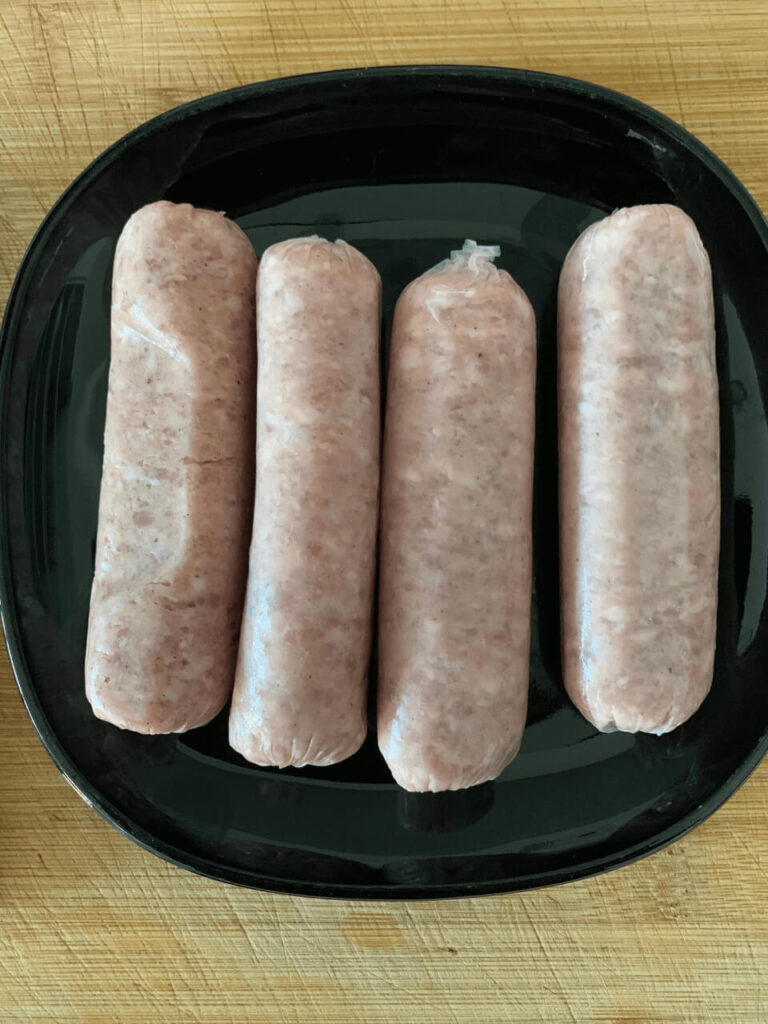 A black plate with four uncooked sausages before being cooked in an air fryer. The sausages are pinkish-white, typical of raw pork, and are neatly placed side by side on the plate.