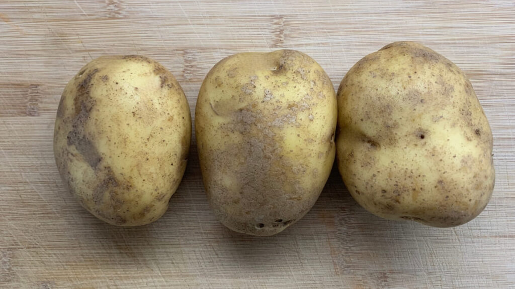 Three unpeeled potatoes with earthy spots are aligned horizontally on a wooden surface, showcasing their natural texture.